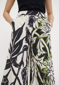 Personne portant un pantalon à jambes larges avec de grands imprimés floraux noirs et verts et les mains dans les poches, associé à un haut noir sans manches.