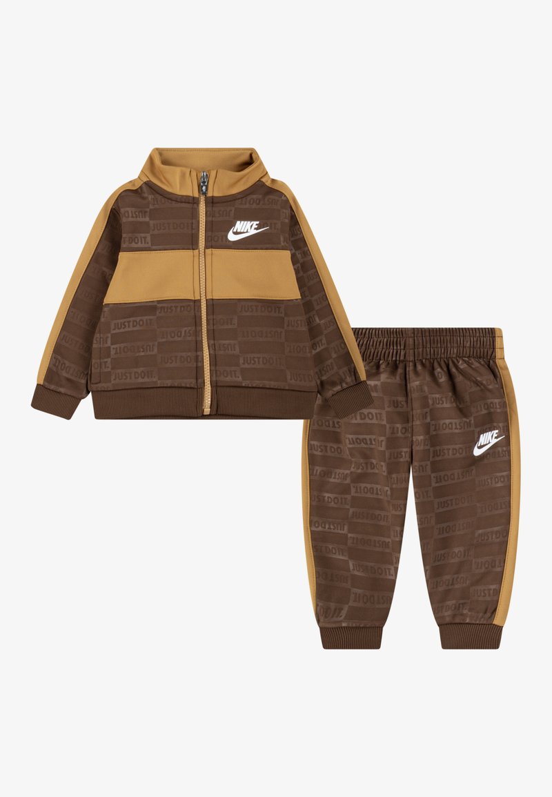 Rjav Nike trenirka, ki vključuje jacketo z zadrgo in elastične hlače. Tekstura blaga s motivom "JUST DO IT" in kontrastnimi peščenimi poudarki. Bela oznaka.