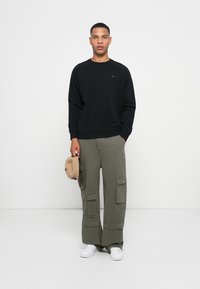 Felpa nera con polsini a costine, abbinata a pantaloni cargo verde oliva con molteplici tasche. Il modello tiene un cappellino beige e indossa sneakers bianche.