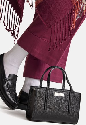 Borsa a mano in pelle nera con texture, con manici corti, posizionata vicino a una gamba in pantaloni borgogna. Calzini bianchi e mocassini neri sono parzialmente visibili.