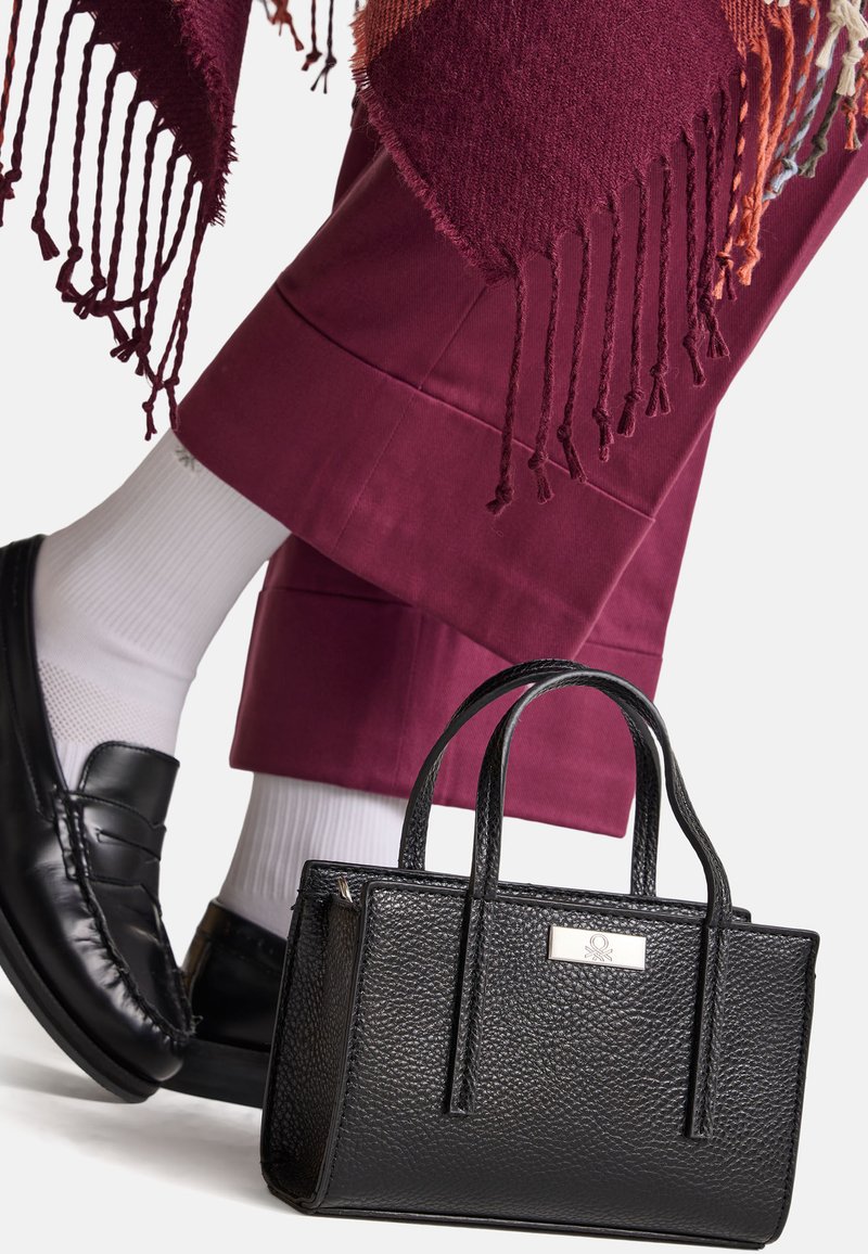 Černá strukturovaná kožená kabelka s krátkými uchy, umístěná poblíž burgundského nohavice. Černé ponožky a černé mokasíny jsou částečně viditelné.