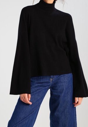 Persoon draagt een zwarte coltrui met lange mouwen en wijde mouwuiteinden, gecombineerd met blauwe, hooggetailleerde jeans tegen een neutrale achtergrond.