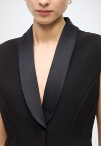 Black garment with a deep V-neck and wide satin lapels. Sleek, structured design emphasizes clean lines and tailored fit.