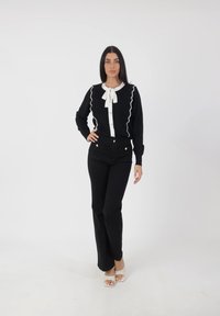 Suéter negro con mangas acanaladas, adornado con un ribete blanco ondulado y un lazo en el cuello, combinado con pantalones negros de cintura alta y sandalias transparentes con tiras.