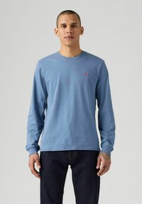 Niebieska bawełniana koszula z długim rękawem, z okrągłym dekoltem i małą czerwoną naszywką na piersi, w zestawie z ciemnymi dżinsami.