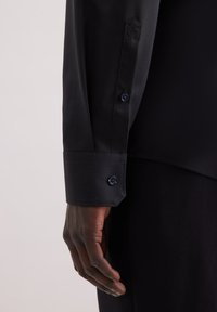 Maglietta nera a maniche lunghe con polsino abbottonato. Presenta una texture liscia e una forma del polsino esagonale, accentuata da bottoni blu navy.