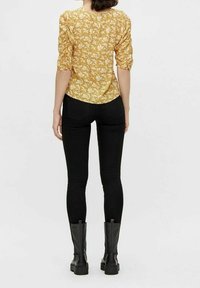 Blouse à manches longues jaune avec motif floral blanc, associée à un jean skinny noir et des bottes noires hautes jusqu'au genou. Texture de tissu lisse notée.