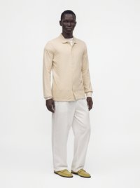 Un homme portant une chemise beige texturée à manches longues, un pantalon blanc ample et des mocassins jaunes, debout devant un fond blanc uni.