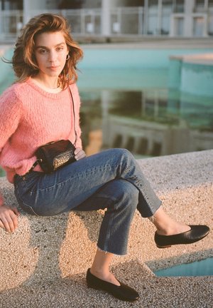 Maglione morbido rosa, jeans blu a vita alta con orli sfrangiati, borsa nera a tracolla, scarpe nere piatte, con acqua turchese sullo sfondo.