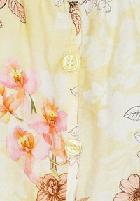 Blomsterbluse med gul baggrund, lyserøde og orange blomster og let tekstur; prydet med runde gule knapper langs fronten.