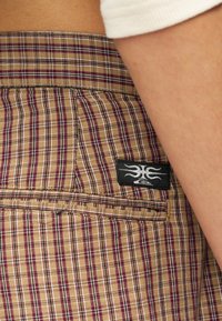 Μπεζ και μοβ καρό υφασμάτινα σορτς με μαύρο και άσπρο κεντημένο λογότυπο κοντά στην πίσω τσέπη, φορεμένα με λευκό τοπ.