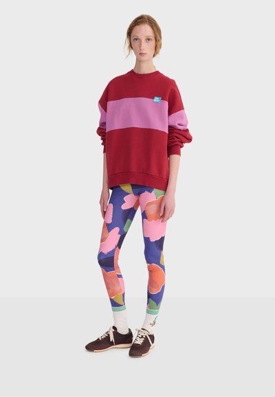Jeune personne debout, portant un sweat-shirt à blocs de couleurs rouge et rose, des leggings floraux multicolores, des chaussettes blanches et des baskets foncées.