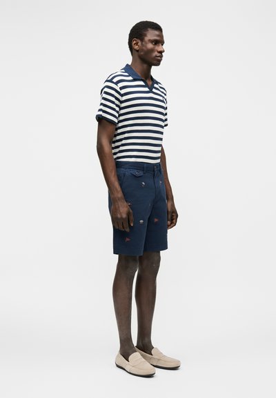 Άνδρας που φοράει μπλε ναυτικό και άσπρη ριγέ πόλο μπλούζα, μπλε ναυτικά σορτς με μικρά κεντημένα σχέδια και μπεζ μοκασίνια σε λευκό φόντο.