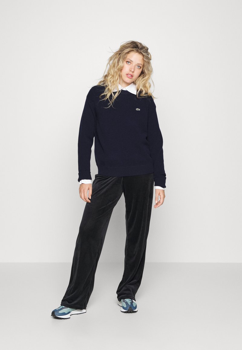 Maglione blu navy con orlo a coste, indossato sopra una camicia bianca con colletto; abbinato a pantaloni in velluto neri e scarpe da ginnastica con dettagli blu.
