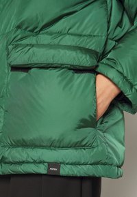 Veste rembourrée verte avec une texture lisse, de grandes poches latérales avec fermetures Velcro et une étiquette AS PESI à l'ourlet.