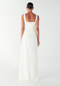 Žena s frizurom u punđu, odjevena u bezrukavnu, bijelu haljinu do poda, stoji okrenuta leđa prema jednostavnoj svijetloj pozadini.