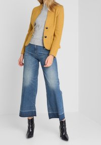 Senfgelber Blazer mit schwarzen Knöpfen, kombiniert mit einem grauen, figurbetonten Pullover und weiten Jeans. Schwarze Stiefeletten runden den Look ab.