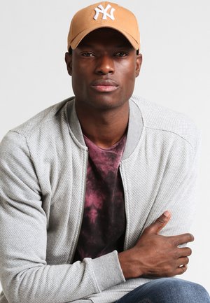 Gray textured bomber jacket with a zip, deep maroon t-shirt underneath, and a tan cap featuring a white logo. Smooth skin, arms crossed.