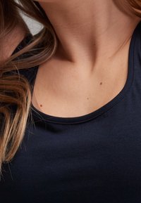 Cou et haut de la poitrine d'une femme à la peau claire, aux cheveux bruns, portant un t-shirt bleu marine foncé à col rond visible.