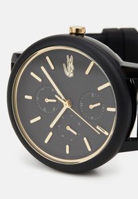 Reloj de pulsera negro con acabado mate, acentos dorados, esfera redonda, tres subesferas y un logo de cocodrilo. Presenta marcadores de hora y agujas doradas.