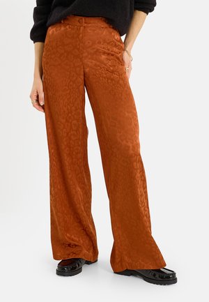 Mujer con pantalones anchos estampados de color óxido, mocasines negros brillantes y un suéter negro, de pie frente a un fondo liso.