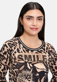 Top à manches longues à imprimé léopard avec un bord noir, comportant le mot "SMILE" en détail clouté et une texture de tissu lisse.