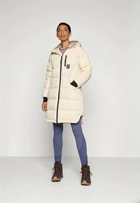 Puhasti kaput u krem boji s kapuljačom, prednjim zatvaračem i crnim detaljima. Nošen preko plavih tajica i smešnih čizama, s logotipom na grudima.