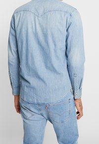 Ljusblå denimskjorta med långa ärmar, spetsskjorta, knäppningar vid ärmslut och sidslitsar. Tillverkad av mjukt, urtvättat tyg med en slät yta.