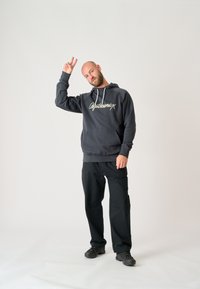 Sudadera gris oscuro con cordones blancos y logo, combinada con pantalones negros y zapatos negros. La persona hace un gesto de paz.