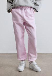 Άτομο που φοράει ανοιχτό ροζ φόρμες, γκρι φούτερ και λευκά αθλητικά παπούτσια, στέκεται σε μπεζ πάτωμα μπροστά σε απλό λευκό φόντο.