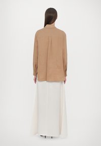 Light brown long-sleeve shirt with a back pleat, paired with a white, floor-length skirt. Smooth fabric with a relaxed silhouette.