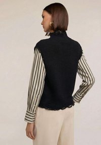 Svart ärmlös stickad väst med hög krage och vågig kant, bärs över en beige och vit randig långärmad skjorta, med en avslappnad passform.