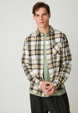 Jonge man in een beige, witte en grijze geruite blouse over een groen T-shirt, staande tegen een effen lichte achtergrond, handen losjes gevouwen.