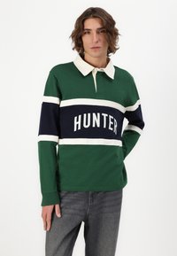 Grønn langermet polo skjorte med marineblå og hvite horisontale striper. Har en hvit krage og "HUNTER"-tekst i fet skrift på brystet.