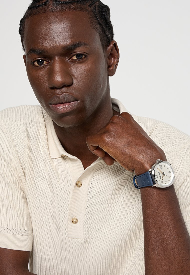 Polo shirt lavorato a maglia beige con maniche corte e tre bottoni, abbinato a un orologio argento con cinturino blu e quadrante chiaro.