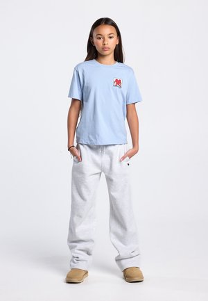 Light blue cotton t-shirt with short sleeves and a square red logo on the chest, paired with gray sweatpants and tan footwear.