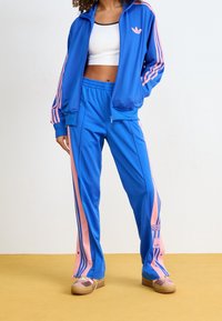 Persoon in een blauwe en roze Adidas trainingsjack en -broek met een witte crop top en roze-bruin sneakers staat op een gele vloer.