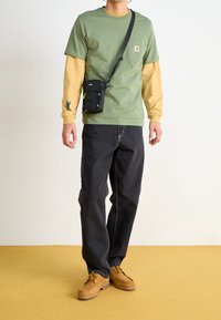 Hombre vestido con camisetas de manga larga verdes y amarillas en capas, jeans negros, botas color topo y una pequeña bolsa de crossbody negra, de pie sobre un suelo amarillo.