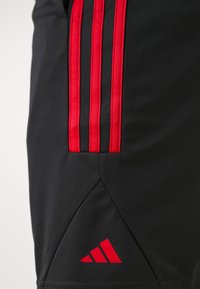 Mustat urheilushortsit, joissa on kolme pystysuoraa punaista viivaa ja punainen Adidas-logo helman lähellä.