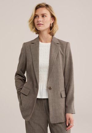 Kvinde iført taupe ternet blazer og matchende bukser med hvidt tekstureret top, stående mod neutral baggrund, hånd i lommen.