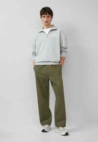 Felpa grigia con zip e un piccolo logo, abbinata a pantaloni olive green a vestibilità rilassata. Il modello indossa scarpe da ginnastica chiare, con le mani nelle tasche.