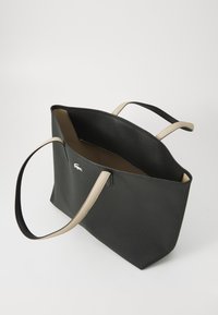 Sac fourre-tout noir texturé avec des poignées beiges et un intérieur beige. Présente un logo de marque bien en vue sur le devant. Design ouvert en haut.