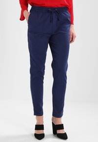 Tmavě modré tepláky s elastickým pasem, šňůrkovým zapínáním a dvěma předními kapsami. Stretchový materiál, úzký střih.