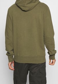 Olivově zelená mikina s kapucí v uvolněném střihu, nastavitelnou kapucí a žebrovanými manžetami a lemem. Materiál působí měkce s minimální texturou.