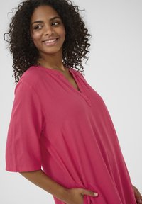 Fuchsia knee-length tunic with a V-neck, short sleeves, and side pockets. Soft, lightweight fabric with a relaxed fit.