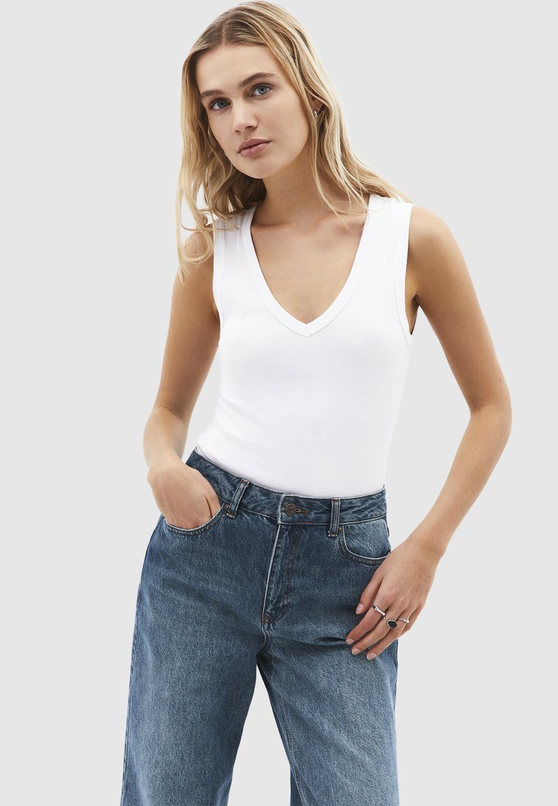 Giovane donna con indosso una maglietta bianca senza maniche a V e jeans blu a vita alta, con una mano in tasca e l'altra appoggiata sulla coscia.