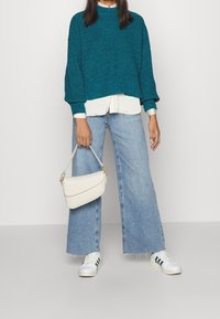 Modrý svetr z pleteniny, pod ním bílá košile s límečkem, světle modré široké džíny, bílá kabelka a bílé tenisky se zelenými pruhy.