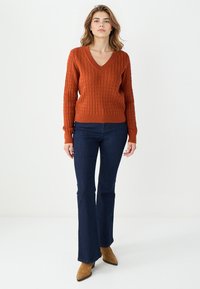 Pull à mailles torsadées couleur rouille avec un col en V et des poignets côtelés, associé à un jean foncé évasé et des bottes à talons marron.