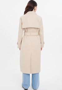 Beige trenchcoat med bälte, bakflik och långa ärmar. Bärs över ljusblå vida jeans och har en slät textur.