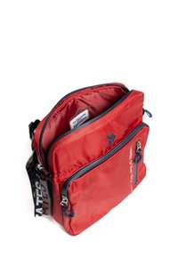 Červená kabelka přes rameno s námořnicky modrými zipy, přední kapsou, částečně otevřeným hlavním přihrádkou a nastavitelným černým popruhem s bílým textem.